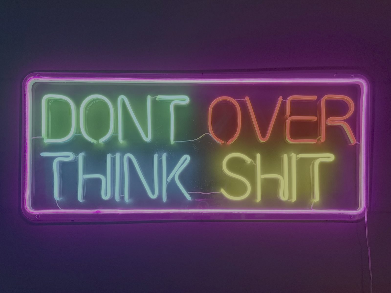 A multicolor neon sign reading DON'T OVER THINK SHIT glowing against a purple wall